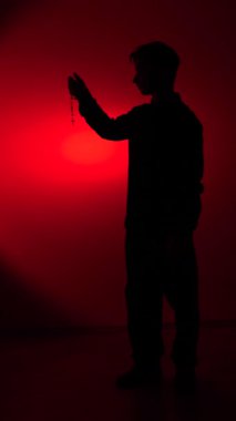 vertical video Man holding rosary beads and praying, his silhouette illuminated by a red spotlight, representing faith, spirituality, and hope in a moment of devotion