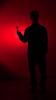 vertical video Persons dark silhouette standing against a vibrant red background, holding a small cross, symbolizing faith, religion, christianity, and spiritual devotion