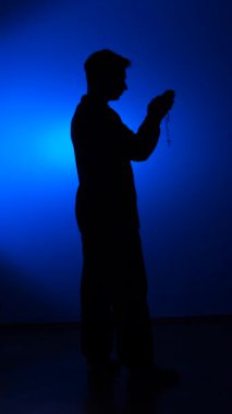 vertical video Silhouette of a man standing in profile, holding rosary beads in clasped hands and praying with devotion against a vibrant blue background with a spotlight effect