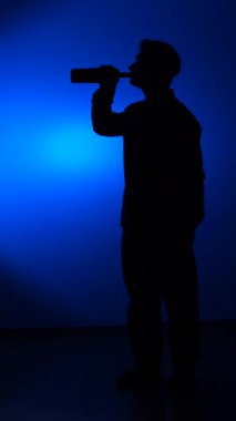 vertical video Man silhouette standing against a blue background, drinking alcohol directly from a bottle, illustrating themes of alcoholism, struggle, loneliness, and addiction