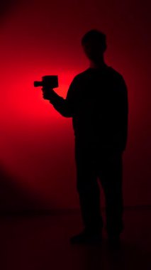 vertical video Silhouette of a person holding a retro film camera, capturing footage against a red background with a dramatic spotlight, representing concepts of filmmaking, nostalgia, and creativity