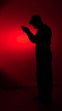 vertical video priest holding rosary beads and praying, his silhouette illuminated by a red spotlight, representing faith, spirituality, and hope in a moment of devotion