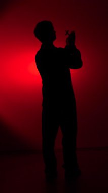 vertical video Silhouette of a man holding a cross and praying with clasped hands, lit by dramatic red studio light against a dark background, symbolizing faith, hope, and devotion