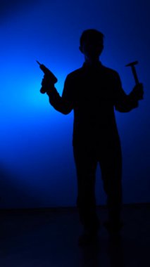 vertical video Man silhouette holding a power drill and hammer, representing construction, repair, handyman work, and diy projects, set against a vibrant blue studio background