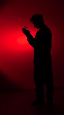vertical video Silhouette of a devout man holding a small cross in his hands, deeply engaged in prayer and reflection, symbolizing faith, devotion, and spirituality against a dramatic red backdrop