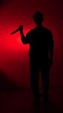 Vertical video mans silhouette standing against a dark red background, holding a sharp knife, evoking concepts of crime, danger, violence, fear, and horror