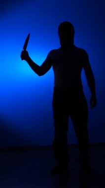vertical video Masked man holding a knife, creating a dark silhouette against a striking gradient blue background, symbolizing danger, threat, crime, and violence with a sense of mystery
