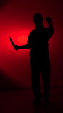 Vertical video male silhouette holding a sharp knife in a threatening manner, standing ominously against a stark red background, conveying danger and fear