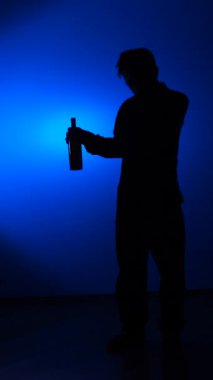 vertical video Person silhouette standing against a blue spotlight background, holding a bottle and depicting concepts of solitude, mystery, addiction, and personal struggles