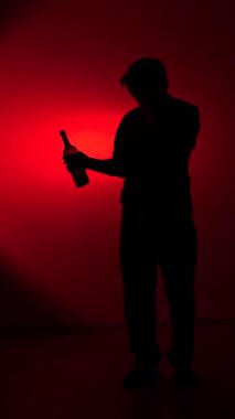 vertical video Man presenting a wine bottle while standing darkly silhouetted against a moody spotlight creating an intense red background, evoking concepts of loneliness and addiction