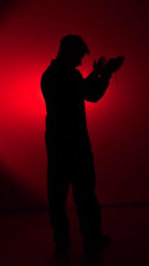 vertical video Dark silhouette of a man bowing in prayer against a red studio background, hands clasped around a rosary, conveying devotion, solitude, and contemplative spiritual connection