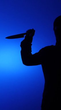 Bıçak tutan bir kişinin dikey video silueti, şiddetli bir tehdit ya da suç eylemi olduğunu gösteriyor, korku, tehlike ve kimliği belirsiz bir saldırganı mavi bir arka plana taşıyor.