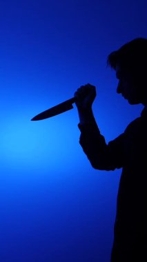 Canlı, mavi bir arkaplana karşı elinde büyük bir bıçak tutan bir kişinin dikey video silueti, tehlikeyi, suçu ve tehdidi karanlık, gizemli bir ortamda taşıyor.