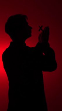 vertical video Silhouette of a man holding a cross and praying with clasped hands, lit by dramatic red studio light against a dark background, symbolizing faith, hope, and devotion