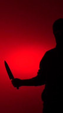 Keskin bir bıçak tutan tehditkar bir adamın dikey video silueti canlı kırmızı bir arka plana karşı ürpertici bir atmosfer yaratıyor tehlike, korku ve korku yayıyor.