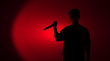 Kırmızı ışıklı arka planda duran gölgeli şahıs, keskin bir bıçak tutuyor, suç, cinayet ve tehdidi temsil eden korkutucu ve tehlikeli bir atmosfer yaratıyor.