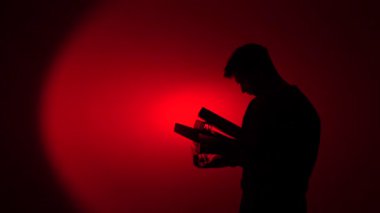 Elinde birden fazla kitap tutan genç bir adamın silüeti, klasör, okuma, eğitim ve dramatik bir kırmızı arkaplan karşısında bilgi arayışı ile derin bir ilişki yansıtıyor.