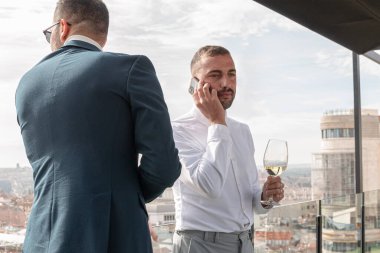 Takım elbiseli iki adam beyaz şarap içerken biri cep telefonuyla konuşuyor. Dışarıda özel bir olayı kutluyorlar..