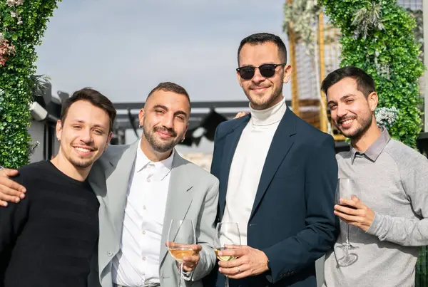Four men friendship. Group of men smiling and holding wine glasses on wedding day, some in casual clothes and others in suits. Outdoors.
