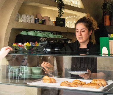 Küçük bir pastanede çalışan bir kadın müşteriye pencereden vejetaryen kruvasan ikram ediyor..