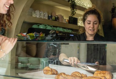 Bayan barista, vejetaryen kahvecide kruvasan servis ederken müşteri heyecanla bekliyor. Siyah üniformalı barista pastayı almak için maşa kullanıyor. Fırın vitrini dolu.