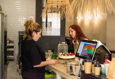 Siyah üniformalı bir barista, samimi bir vejetaryen kahve dükkanında müşteriye hizmet eder. Tezgahta güzel dekore edilmiş bir pasta, kahve fincanları ve dokunmatik ekran POS sistemi ile birlikte sergileniyor. Sıcak.