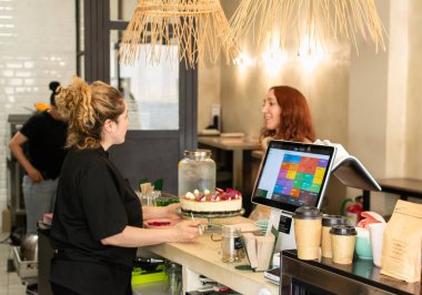 Bir bayan kahve dükkanı sahibi küçük bir işletmenin tezgahında gülümseyen bir müşteriye hizmet eder. Dekorasyonda modern bir satış noktası, sürdürülebilir kahve fincanları ve güzel dekore edilmiş bir pasta yer alıyor.