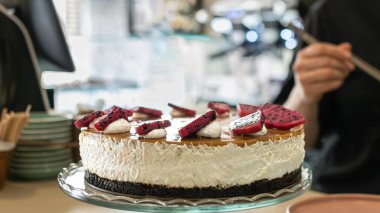 Koyu kurabiye hamurlu, karamelize edilmiş, canlı ejderha meyvesi ve krem şantili yeni hazırlanmış vejetaryen peynirli kekin yakın çekimi. Tatlı cam pasta standına konur.