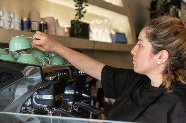 Siyah üniformalı bir garson vejetaryen kahve dükkanındaki profesyonel espresso makinesinden bir fincan yeşil kahve alıyor. Kahve demleniyor. Konforlu ve şık iç mekan sıcak bir atmosfer yaratıyor.