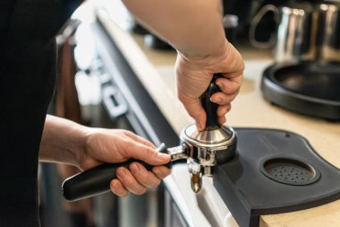 Barista 'nın elinin portatif filtrede öğütülmüş kahveye baskı uyguladığı yakın plan bir fotoğraf. Görüntü, bir uzmanlık alanı dahilinde espresso hazırlığında gerekli teknik ve hassasiyeti vurgular.