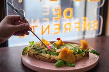 Şef, üzerinde avokado, somon, tofu, lahana ve diğer taze malzemeler bulunan vejetaryen tostunu dikkatlice hazırlamak için cımbız kullanıyor.. 