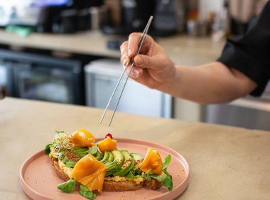 Şef titizlikle vejetaryen avokado tostunu vejetaryen füme somonla süslemek için cımbız kullanıyor.