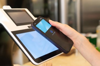 Bir bayan müşteri dijital cüzdanıyla modern bir POS terminalinde akıllı telefon kullanarak ulaşılamaz bir ödeme yapıyor. Görüntü, cep telefonunun kolaylığını ve verimliliğini vurgular