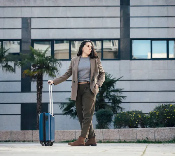 Mavi bir bavul taşıyan, şık giysili genç bir iş kadını, palmiye ağaçları olan modern bir binanın önünde duruyor..