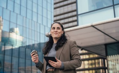 İş kadını elinde bir ofis binasının önünde kahve ve tablet tutarken yukarı bakıyor ve gülümsüyor şehir ortamında dinlendirici bir kahve molası veriyor.