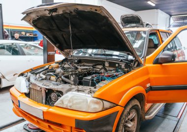 Profesyonel bir araç atölyesinde elektrik sistemi denetimi sırasında kaputu açık, tanısal araçlara ve kablolara bağlı turuncu araba..