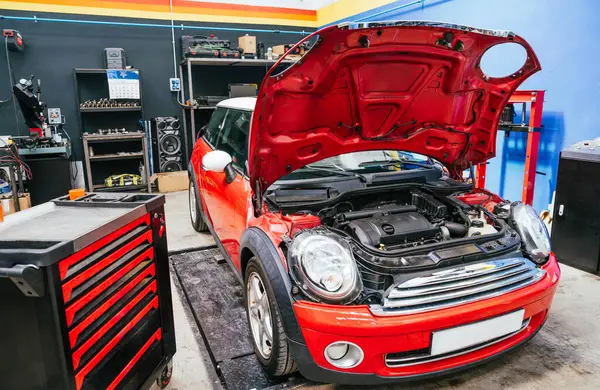 Profesyonel bir atölyede kaputu açık kırmızı bir aracın önü. Etrafı alet ve ekipmanlarla çevrili. Yanındaki, taşınabilir bir çalışma masası. Hassas tanı ve mekanik bakım
