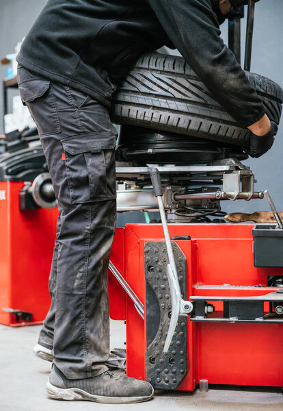 Mechanic prepares to service a car tire, securing it on a red mounting machine. The image highlights setup procedures, physical work, and attention to safety in a real workshop setting.