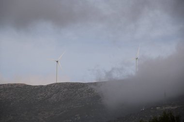 Dağdaki bulutların arkasındaki tepede bir grup rüzgar türbini.