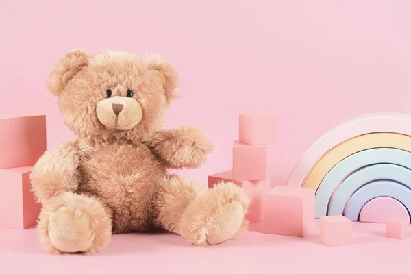Baby kid toys background. Toy teddy bear, pastel color wooden rainbow and pink brick cubes on pastel pink background. Front view.