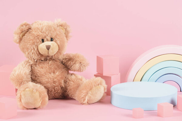 Детские игрушки фон. Пустой круглый стенд подиума и плюшевый мишка, радуга пастельного цвета и деревянные образовательные игрушки на пастельно-розовом фоне. Вид спереди.