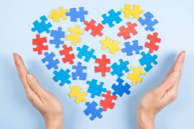 World autism awareness day concept. Woman hands holding colorful puzzle heart on light blue background. Top view, copy space.