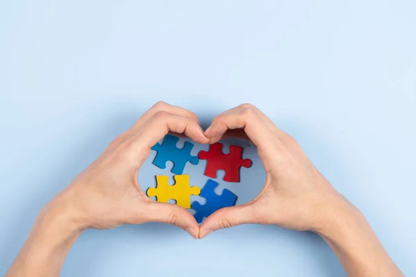 World autism awareness day concept. Woman made heart with her hands over colorful puzzle pieces on light blue background. Top view, copy space.