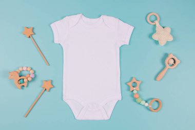Beyaz pamuklu bebek kısa kollu vücut giysisi ve açık mavi arka planda doğal ahşap oyuncak. Bebek tulumu maketi. Boş cinsiyet nötr yeni doğmuş vücut giysisi maketi. Üst görünüm.