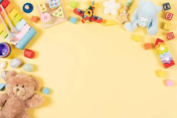 Baby kids toy frame background. Teddy bear, colorful wooden educational, musical, sensory, sorting and stacking toys for children on pastel yellow background. Top view, flat lay.