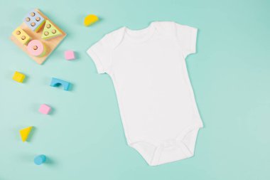 Beyaz pamuklu bebek kısa kollu vücut giysisi ve pastel arka planda doğal ahşap çevre dostu eğitim oyuncakları. Bebek tulumu maketi. Boş cinsiyet nötr yeni doğmuş vücut giysisi maketi. Üst görünüm.