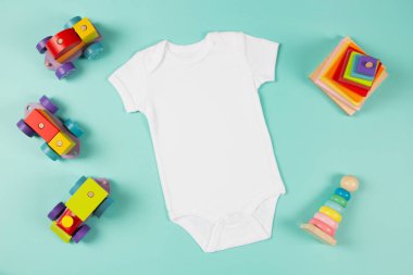 Beyaz pamuklu bebek kısa kollu vücut giysisi ve yeşil arka planda doğal ahşap çevre dostu eğitim oyuncakları. Bebek tulumu maketi. Boş cinsiyet nötr yeni doğmuş vücut giysisi maketi. Üst görünüm.