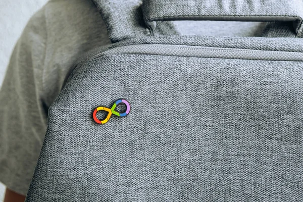 Teenage boy with autism infinity rainbow symbol sign metallic pin brooch on gray backpack. World autism awareness day, autism rights movement, neurodiversity, autistic acceptance movement.