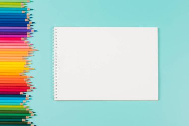 Abstract background for painting, drawing and sketching. Blank white paper spiral notebook and many colored pencils on pastel green background. Top view, flat lay, copy space for text.