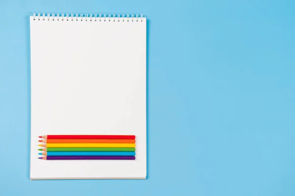 Open white notebooks with purple, blue, green, yellow, orange, red colored pencils on light blue background. Top view, copy space for text.